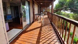 A wooden deck with a glass door leading to a room, a table with chairs, and a view of greenery. - Mar Azul 18 (Ponta d'Ouro)