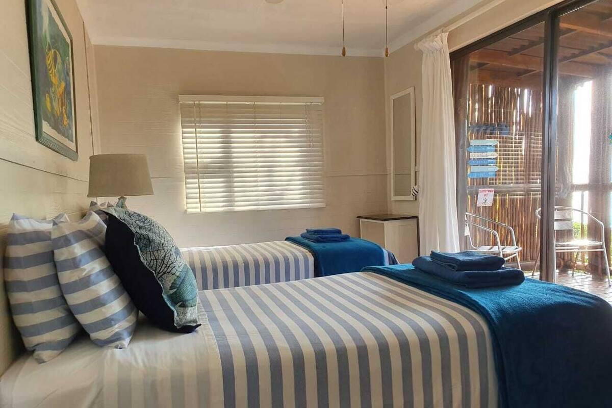 A hotel room with two beds, a window with blinds, a lamp, and a view of an outdoor area with chairs.