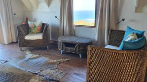 A room with a high ceiling, a large window with curtains, wicker furniture, and a view of the ocean. - Mar Azul 18 (Ponta d'Ouro)