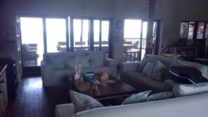 A spacious living area with a large sofa, a coffee table, and a television. There is a balcony visible through the large windows. - Mar Azul 1B (Ponta d'Ouro)