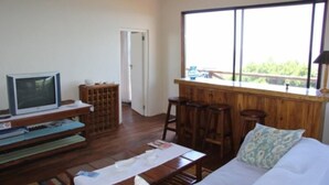 A room with a bed, a TV on a stand, a wooden bar area with stools, and a large window offering a view of greenery. - Mar Azul 26 (Ponta d'Ouro)
