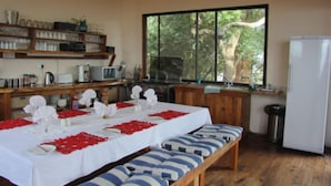 A dining area with a long table set for four, benches, a refrigerator, and a window with a view of trees. - Mar Azul 26 (Ponta d'Ouro)