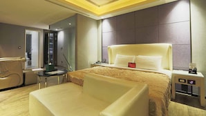 A modern hotel room with a large bed, a sitting area with a chair, a small table, and a bedside table with a lamp.