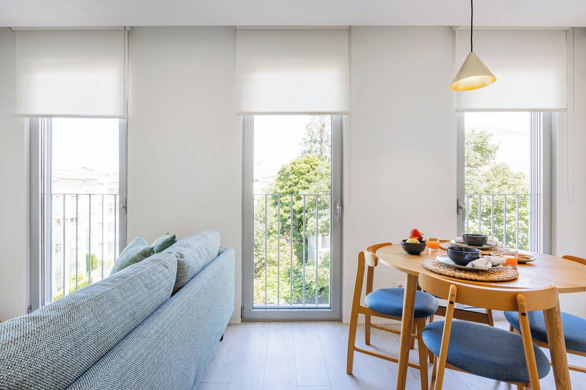 A modern living room with a sofa, dining table, chairs, and a view through large windows.