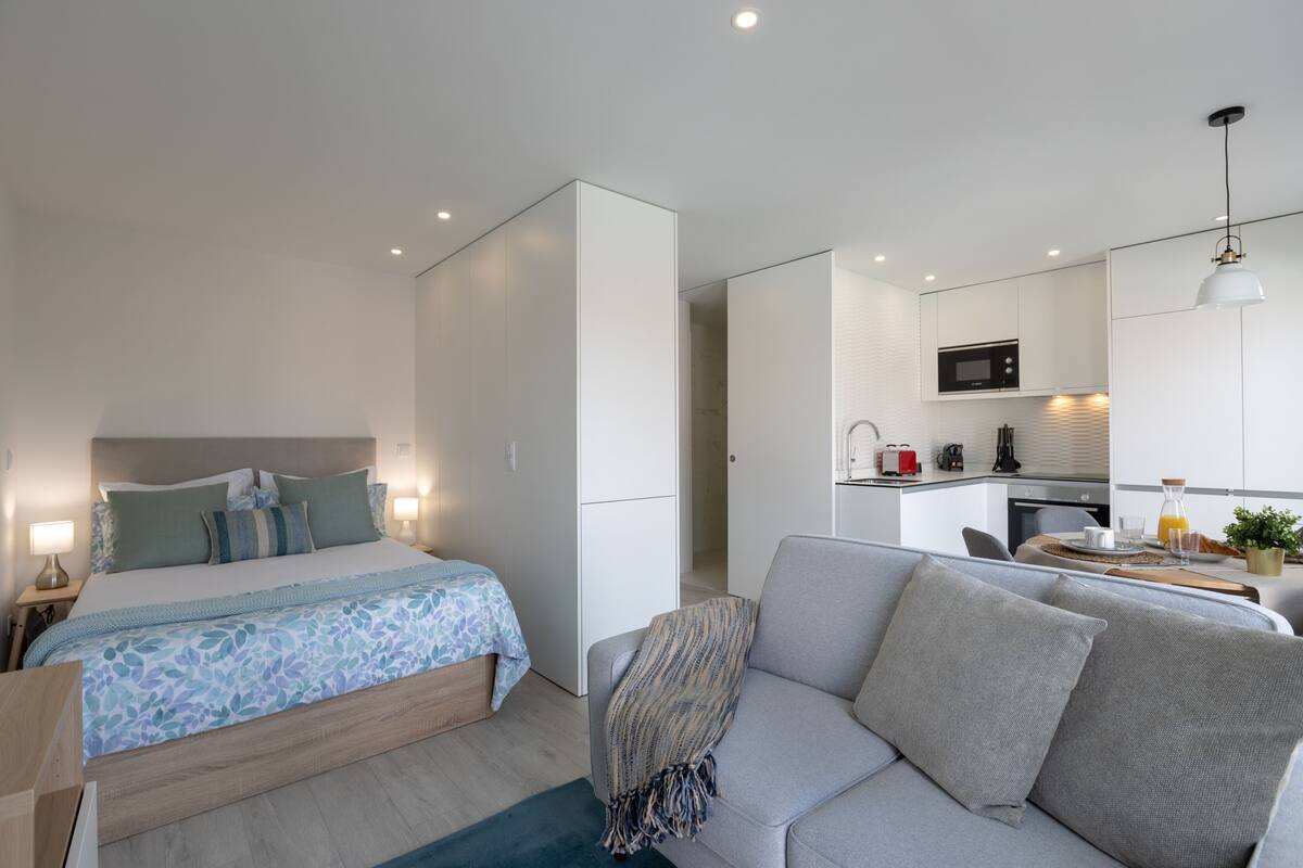A modern hotel room with a bed, a sofa, a kitchenette, and a bathroom.
