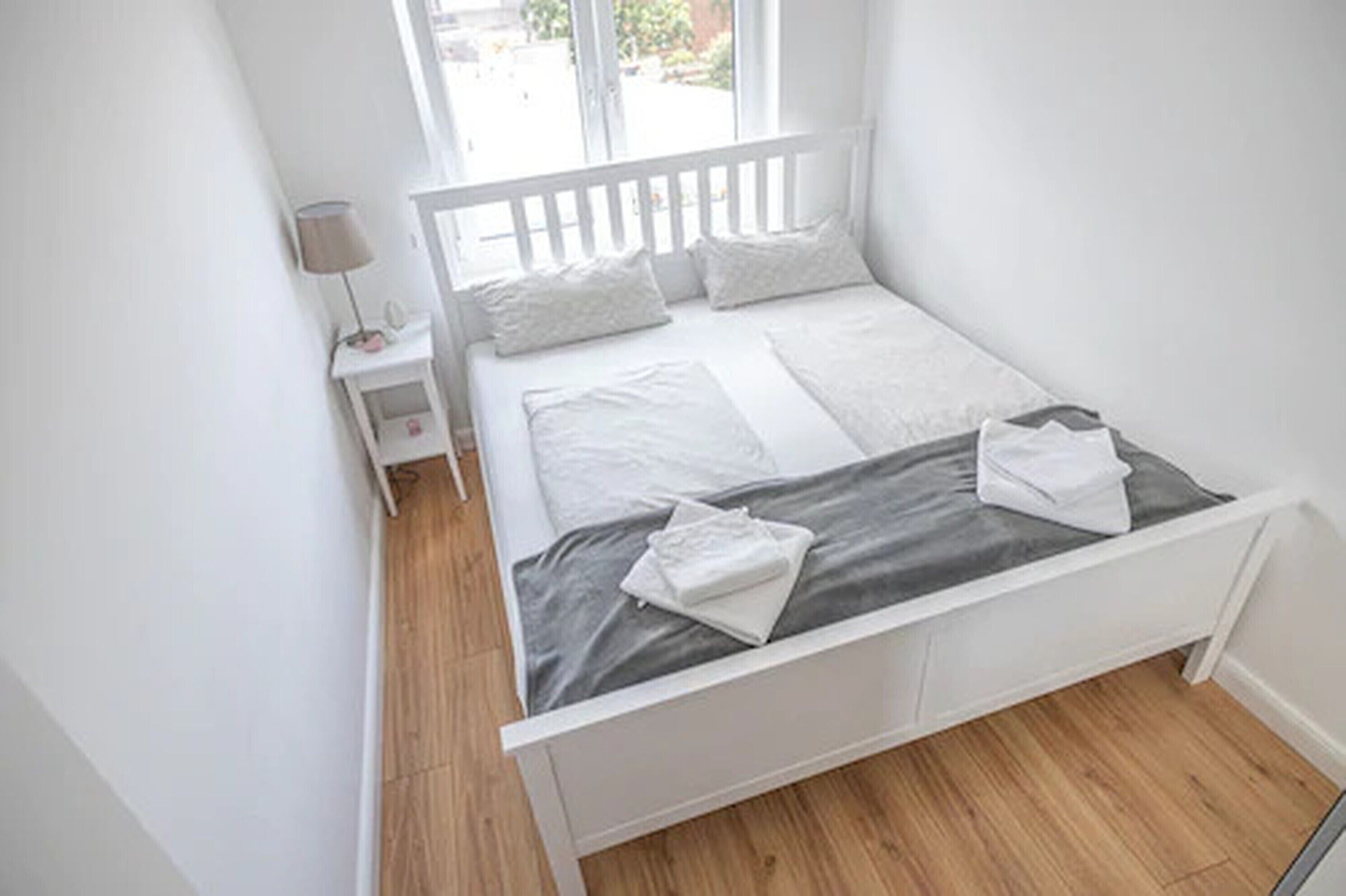 A small, white, single-bed room with a wooden floor, a nightstand, and a window with a view of the outside.
