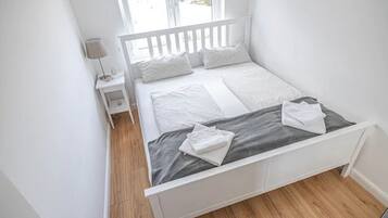A small, white, single-bed room with a wooden floor, a nightstand, and a window with a view of the outside.