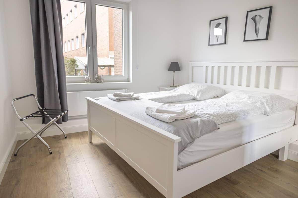 A neatly made bed with white linens, a bedside table with a lamp, and a window with curtains.