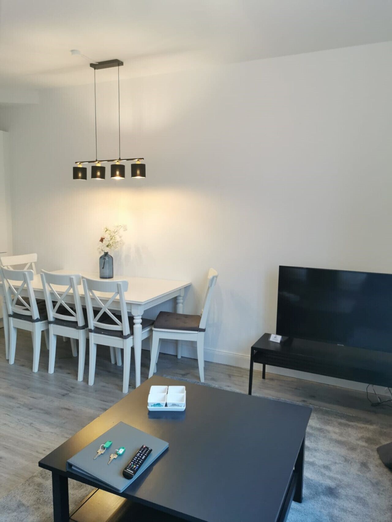 A modern living room with a dining area, a flat-screen TV, and a black coffee table with remote controls and keys.