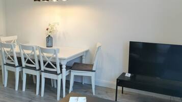 A modern living room with a dining area, a flat-screen TV, and a black coffee table with remote controls and keys.