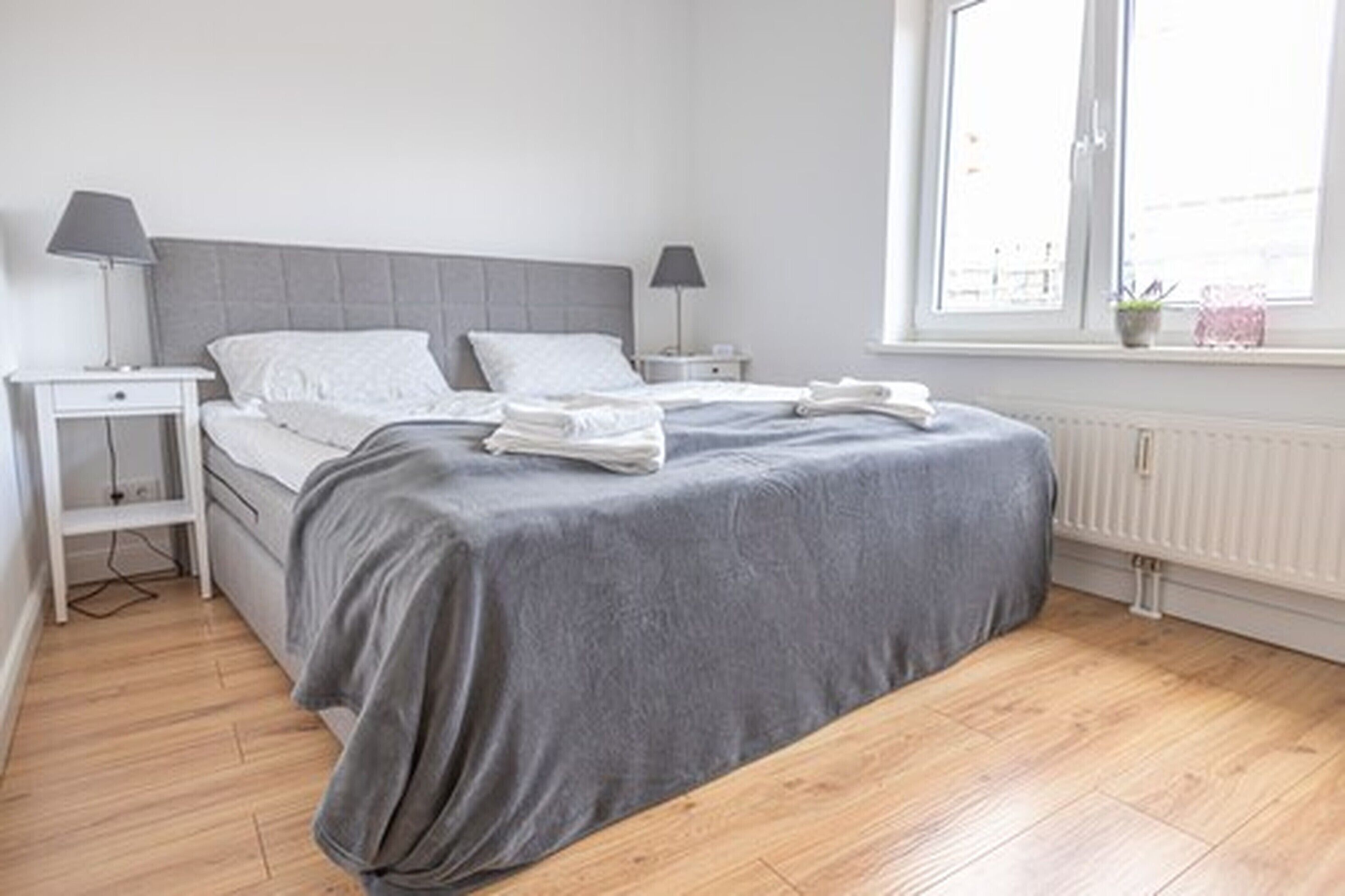 A neatly made bed with a grey bedspread and white pillows, two bedside tables with lamps, a window with a view, and a radiator.