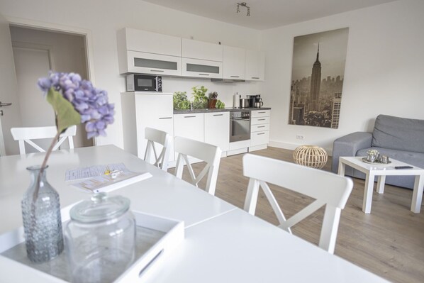 A modern kitchen with white cabinets and a microwave, a dining area with a white table and chairs, and a grey sofa in a living area with a cityscape poster on the wall. - Arbio I Dehnhaide Apartments (Hamburg)