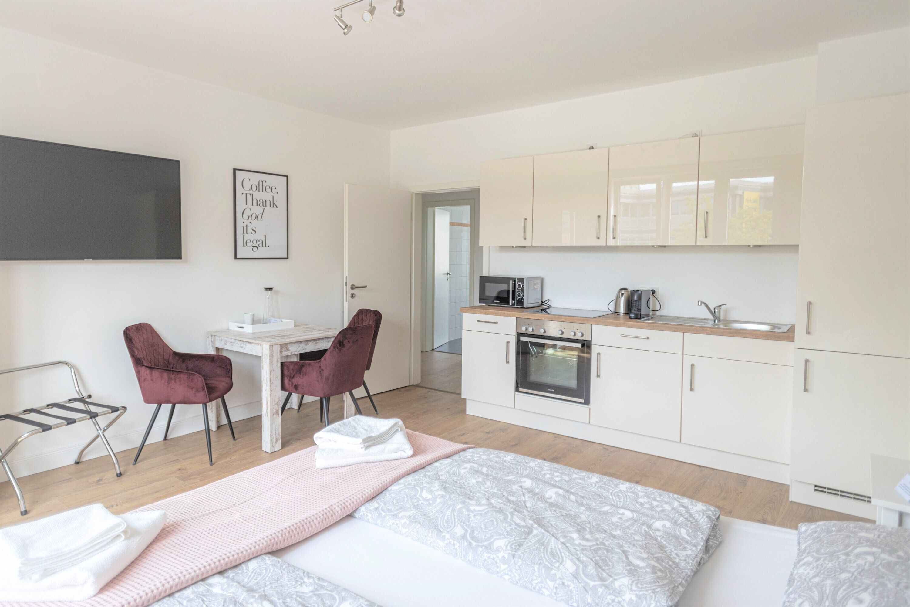 A modern, compact room with a bed, a small desk, two chairs, a kitchenette, and a TV mounted on the wall.