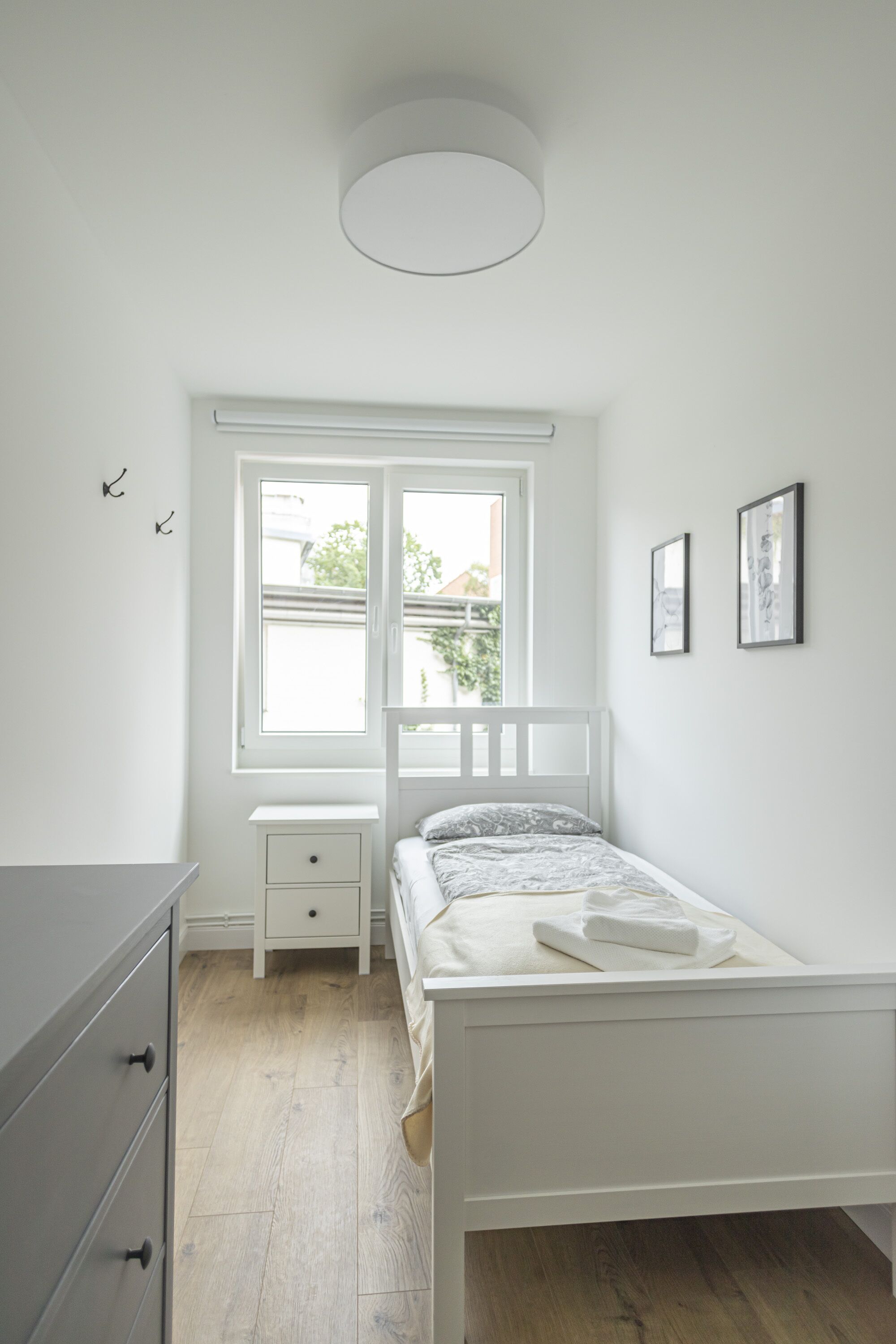 Una camera da letto con un letto bianco, un comodino, una cassettiera e una finestra vista sul verde.