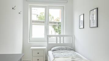 Una camera da letto con un letto bianco, un comodino, una cassettiera e una finestra vista sul verde.