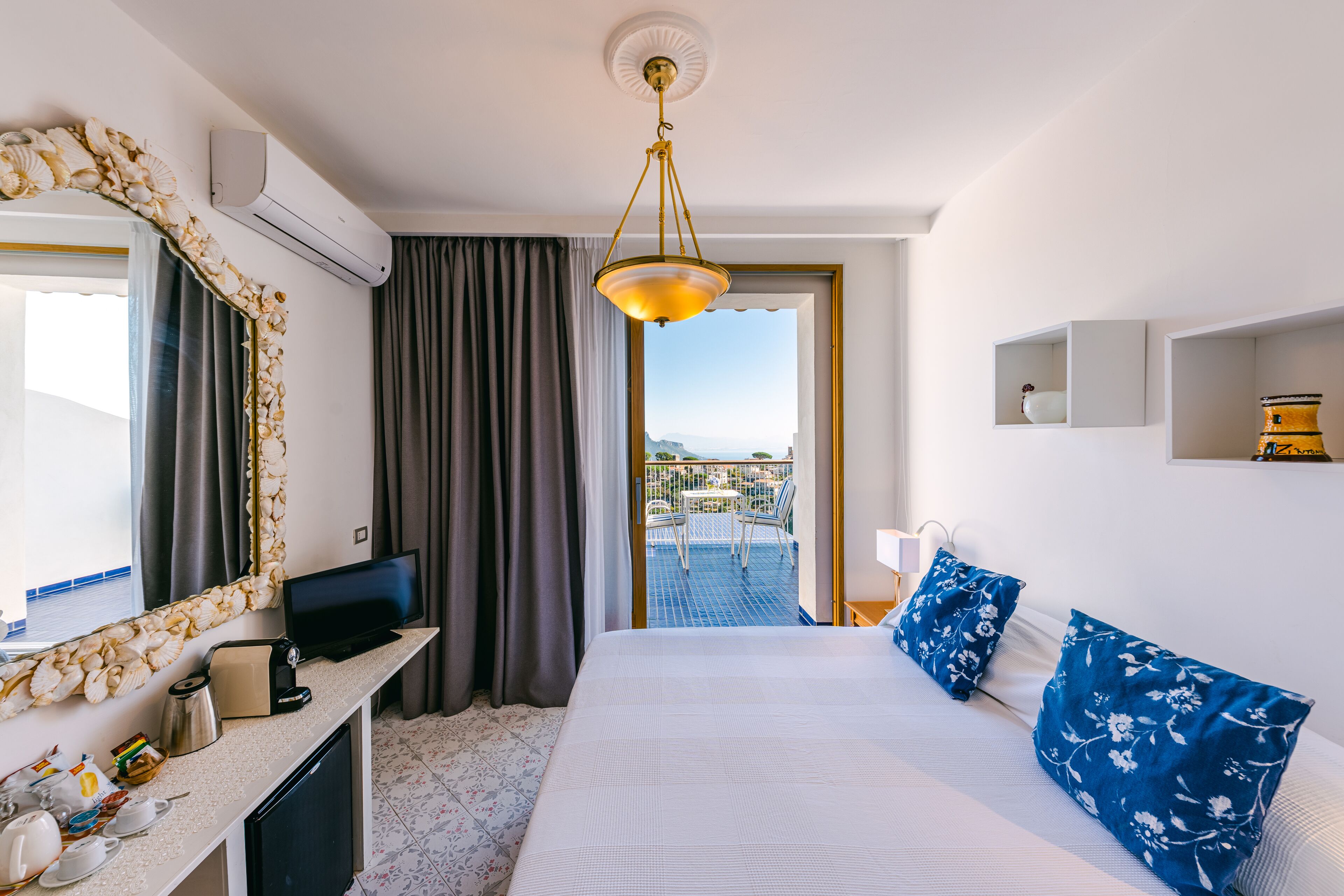 Habitación de hotel con cama, escritorio, televisión, aire acondicionado, balcón con vistas y un espejo decorativo.