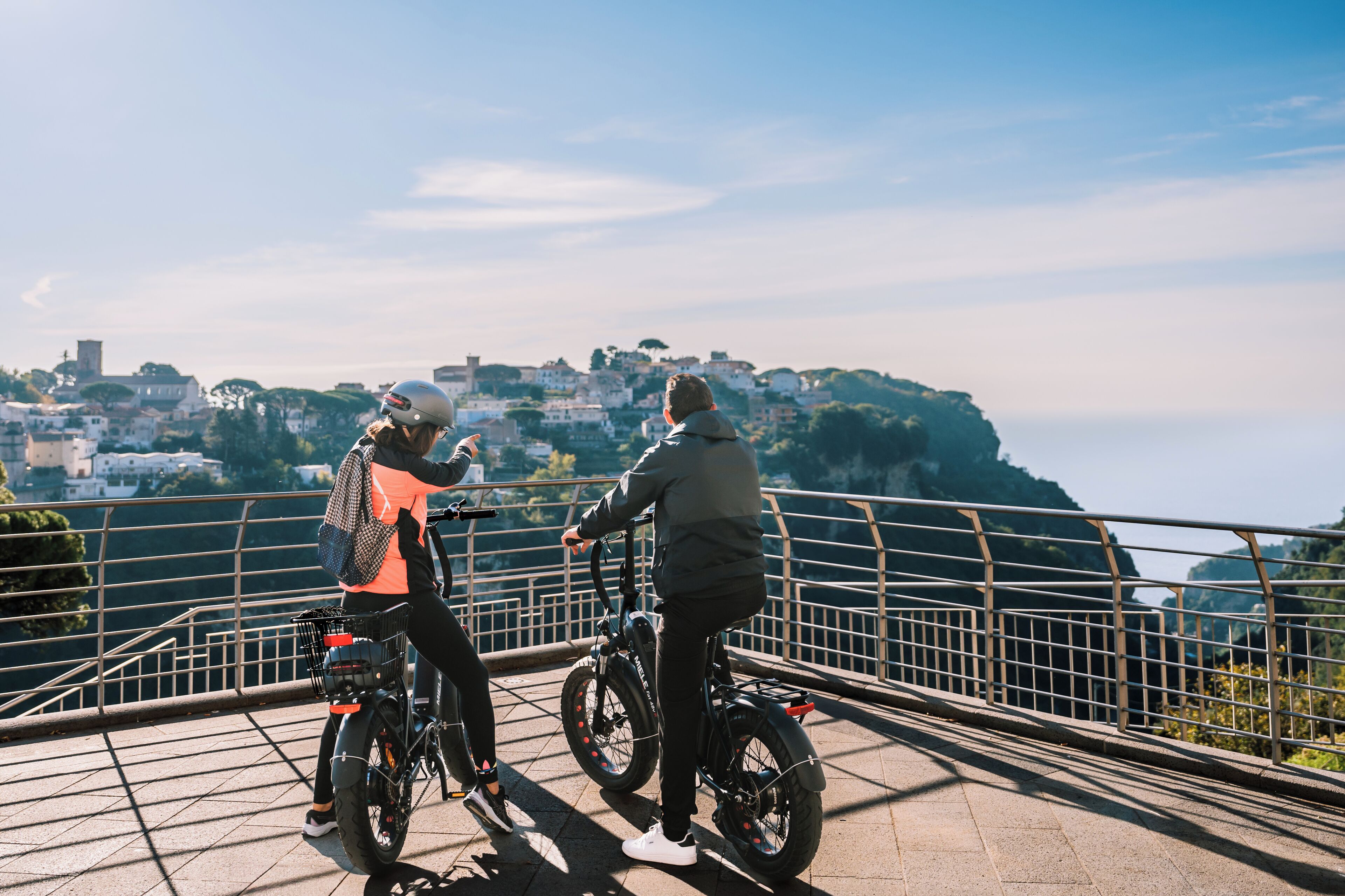 Duas pessoas de bicicleta numa ponte, com vista panorâmica de uma cidade ao fundo.