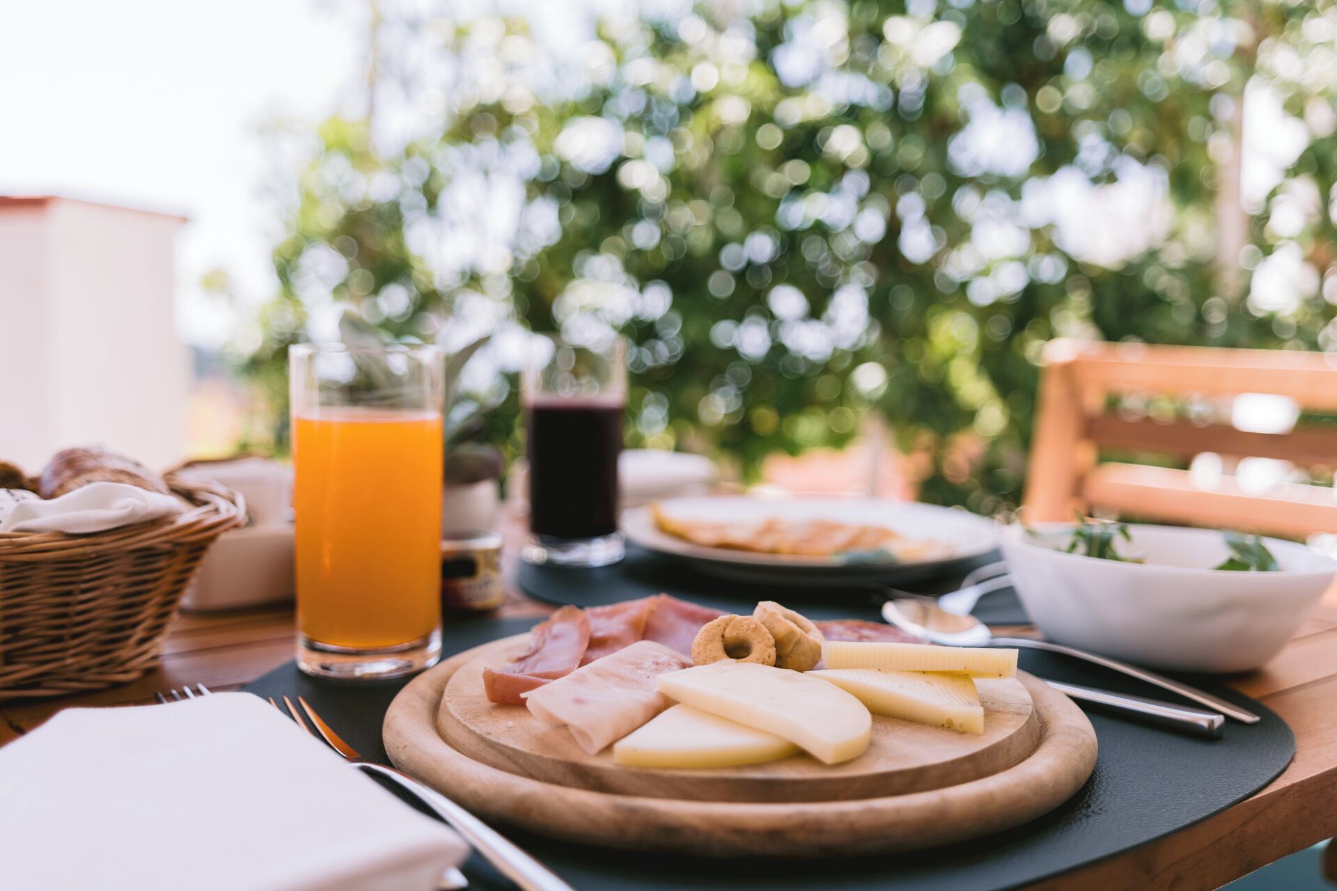 Un desayuno con una tabla de madera de quesos, jamón y pan, un vaso de zumo de naranja y una bebida oscura.