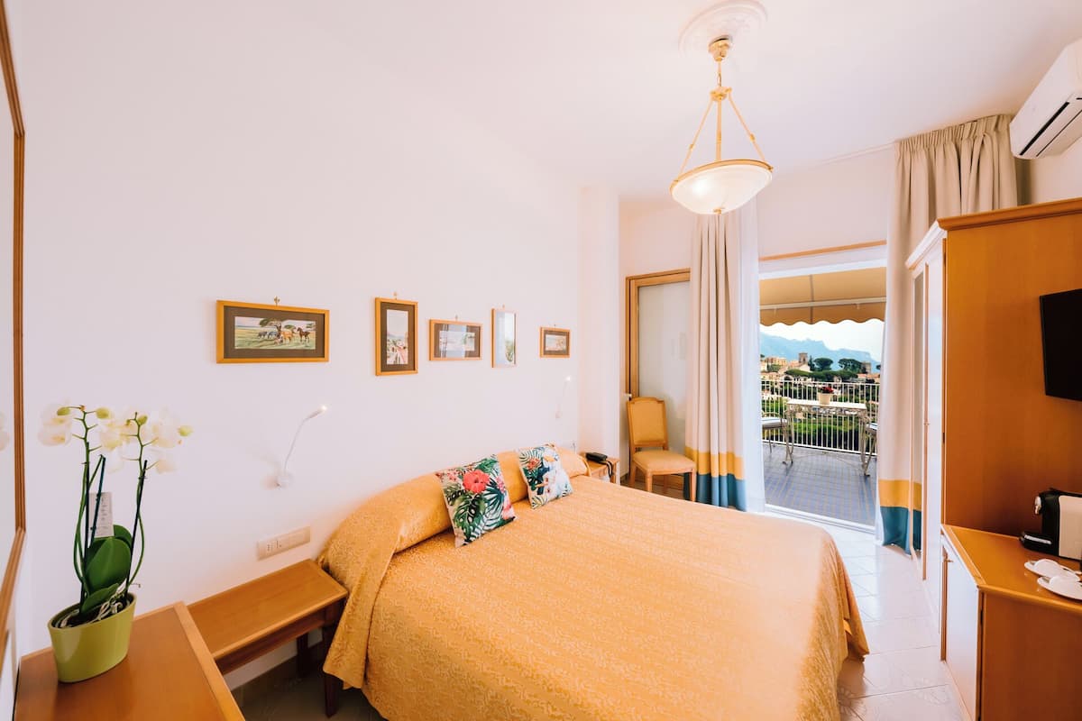 Una habitación de hotel con cama, escritorio, silla, planta en maceta y balcón con vistas.