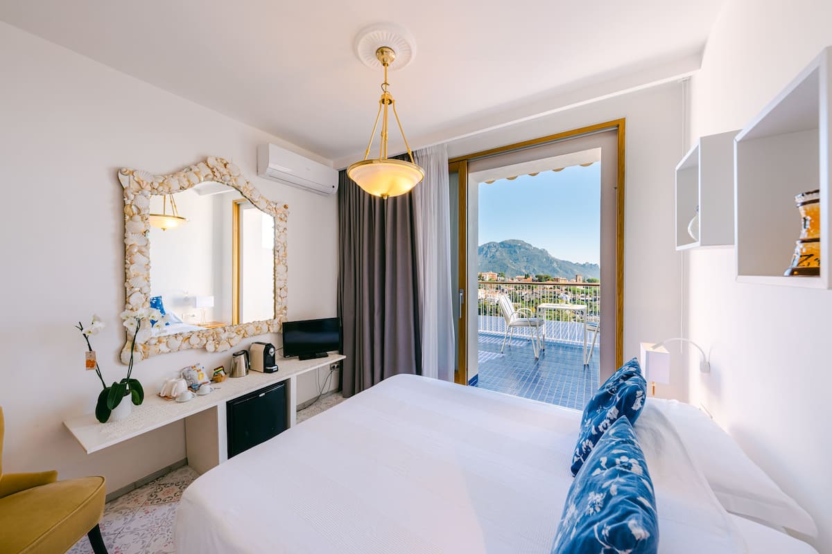 Habitación de hotel con cama, escritorio, televisor, espejo y balcón con vista a las montañas.