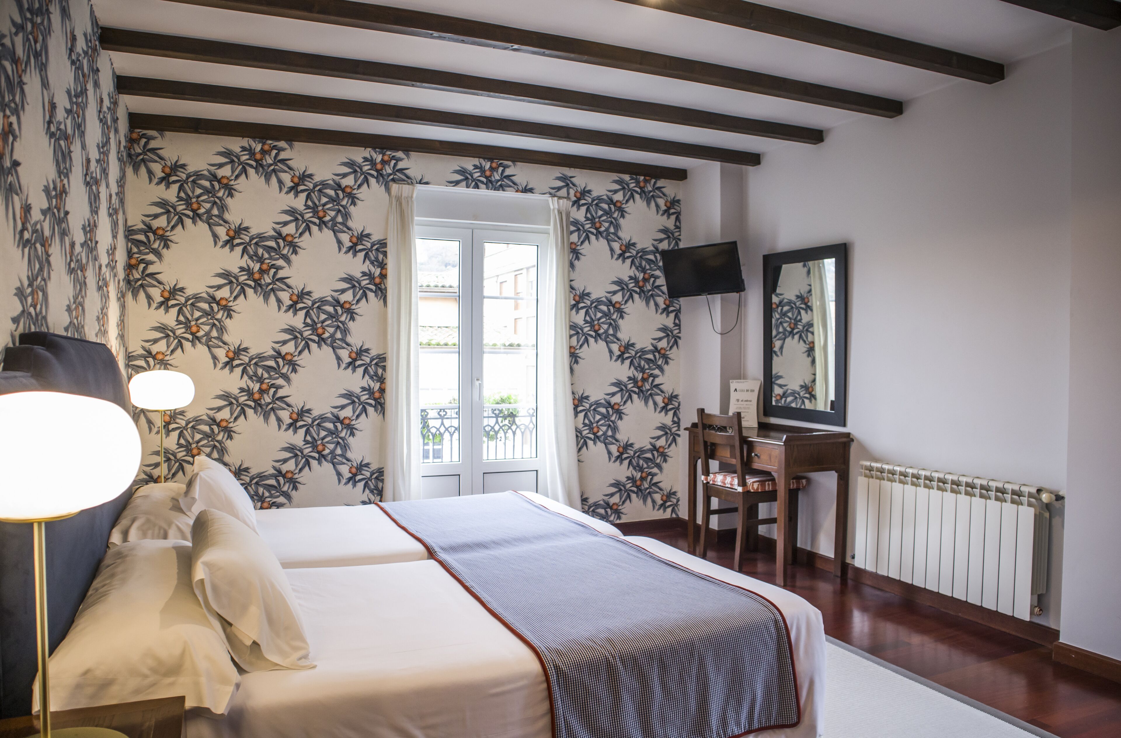 A hotel room with a bed, bedside lamps, a desk, a chair, a TV, and a window with a view of the outside.