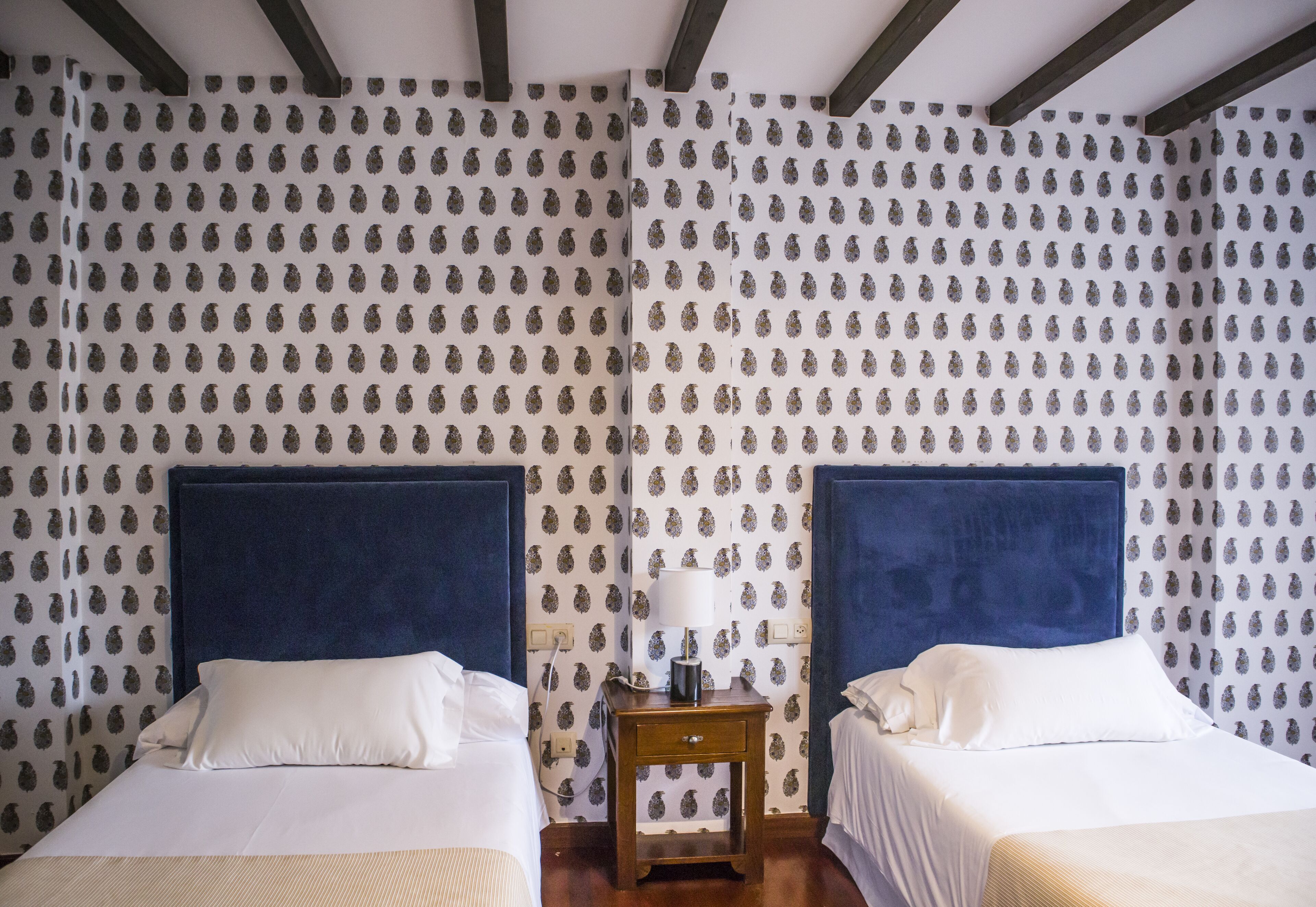Een hotelkamer met twee bedden, een nachtkastje met een lamp, en behang met een patroon.