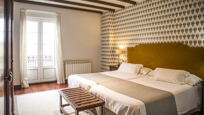 Habitación de hotel con una cama grande, un banco de madera, un papel tapiz estampado y vistas al exterior por una puerta.