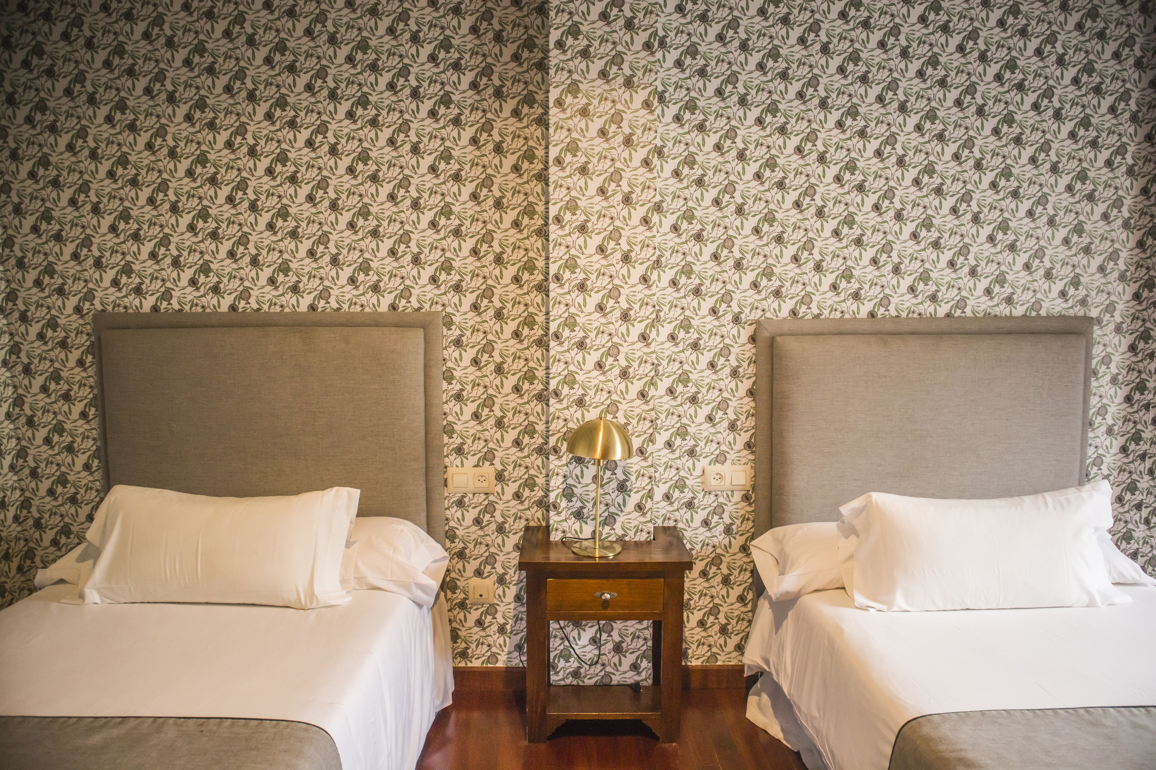 Habitación de hotel con dos camas, una mesita de noche con lámpara y papel tapiz estampado.