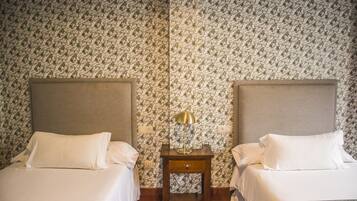 Habitación de hotel con dos camas, una mesita de noche con lámpara y papel tapiz estampado.