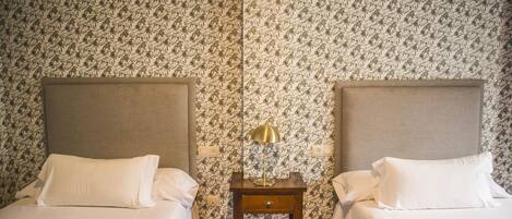 Habitación de hotel con dos camas, una mesita de noche con lámpara y papel tapiz estampado.