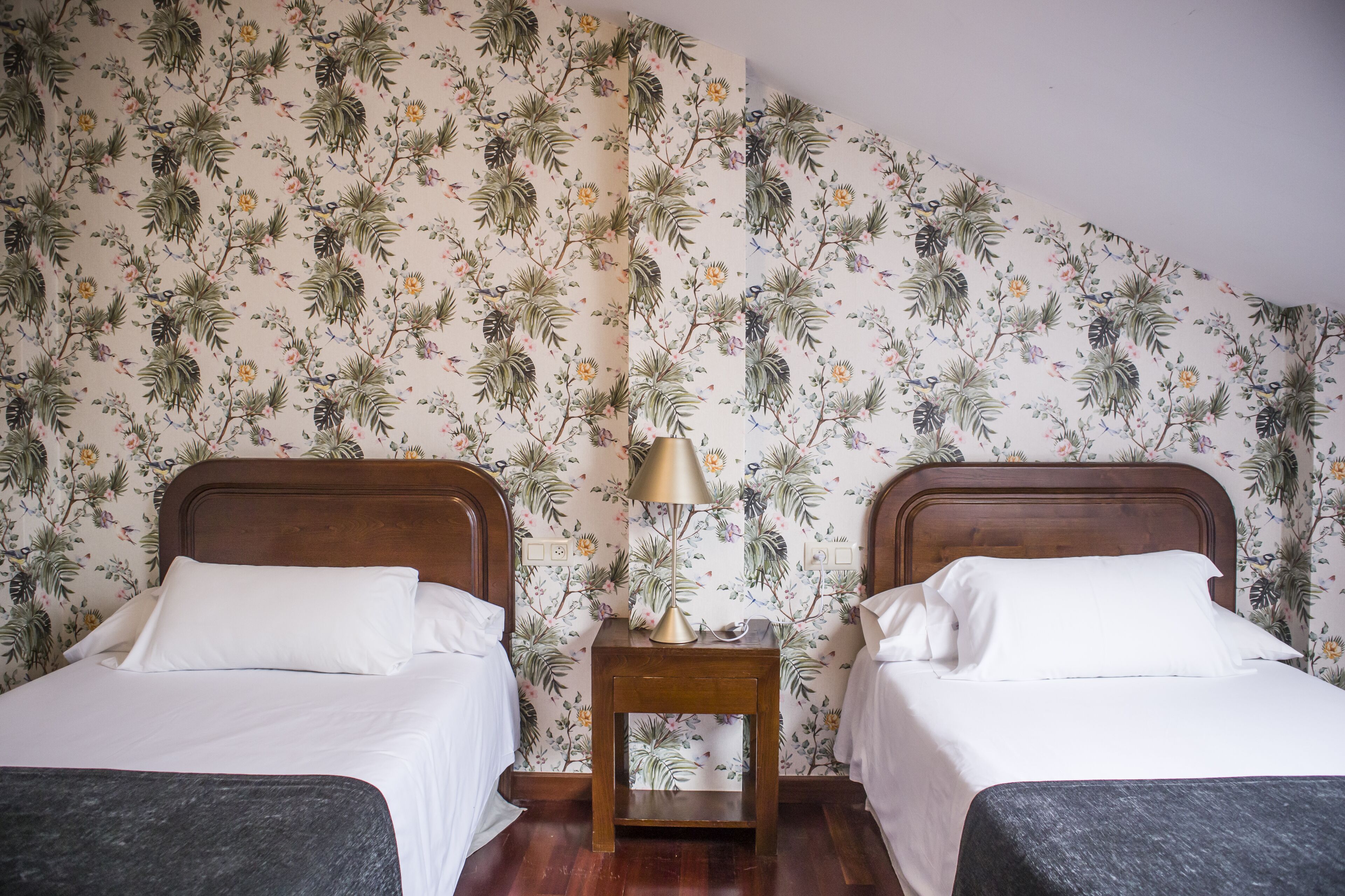 A bedroom with two beds, a nightstand with a lamp, and floral wallpaper.