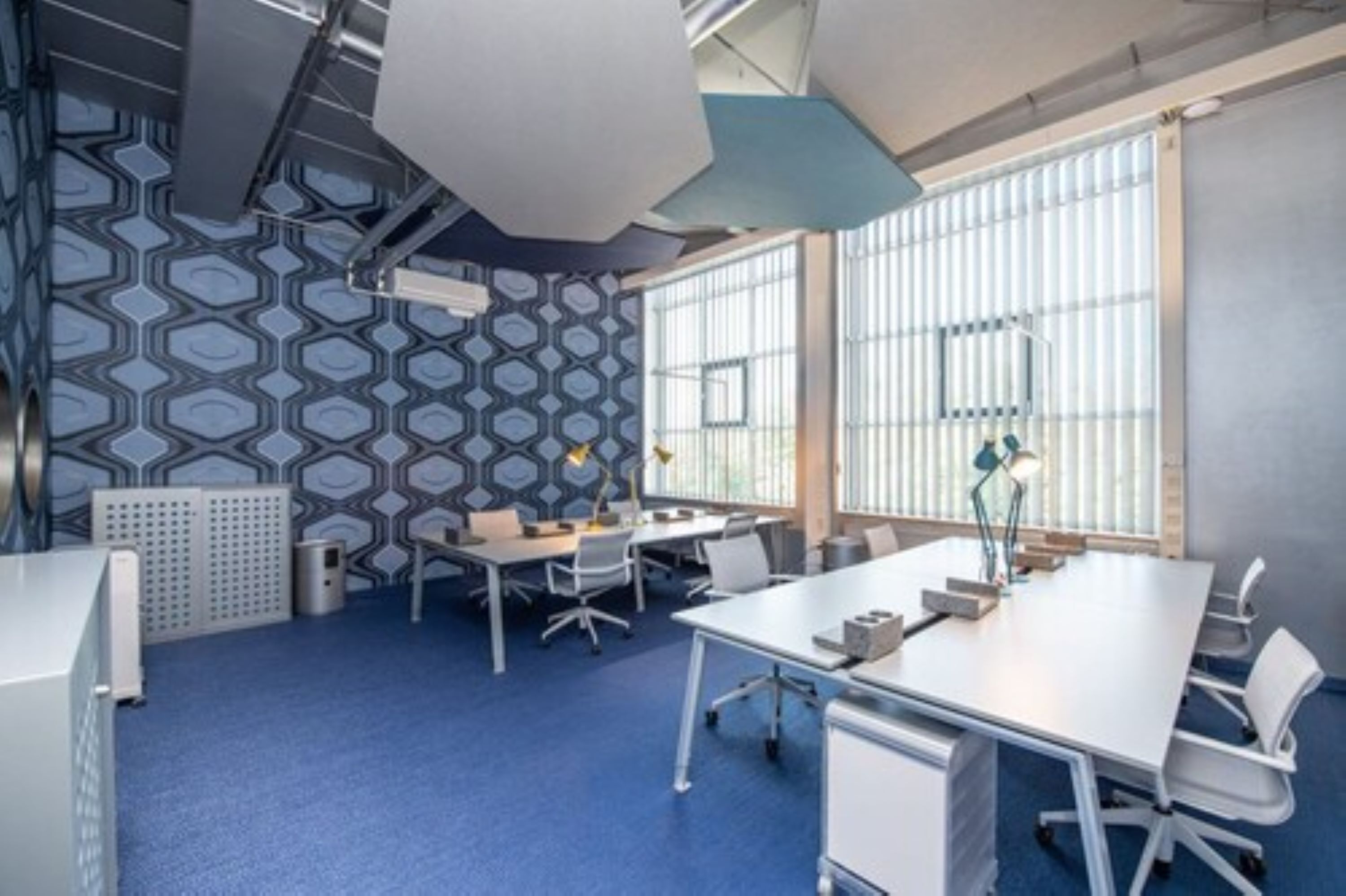 A modern office space with a blue carpet, white desks, and chairs, and large windows with blinds.