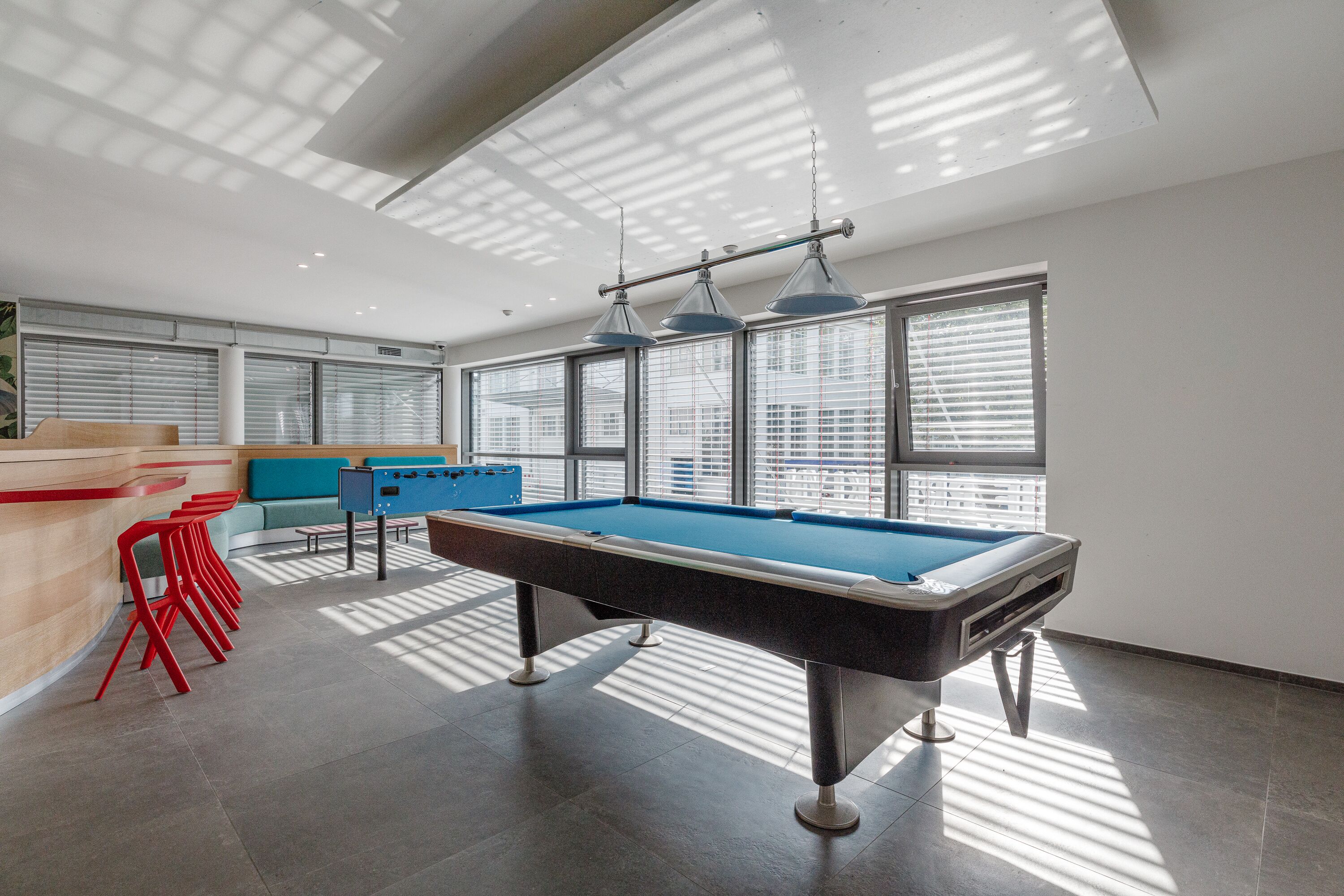 A pool table in a well-lit room with large windows, red chairs, and a blue sofa.