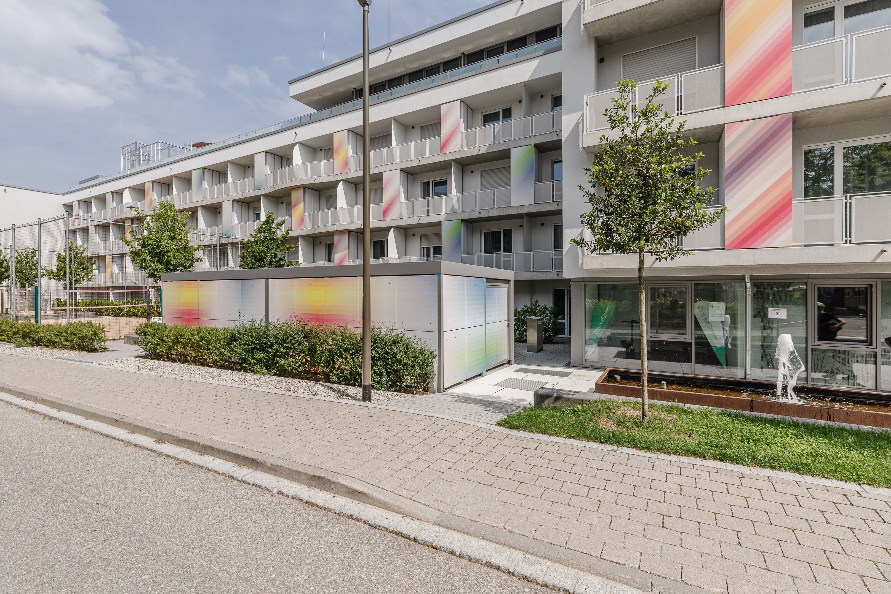 A modern multi-story building with balconies, a glass facade, and a landscaped area with a small tree and a fountain.