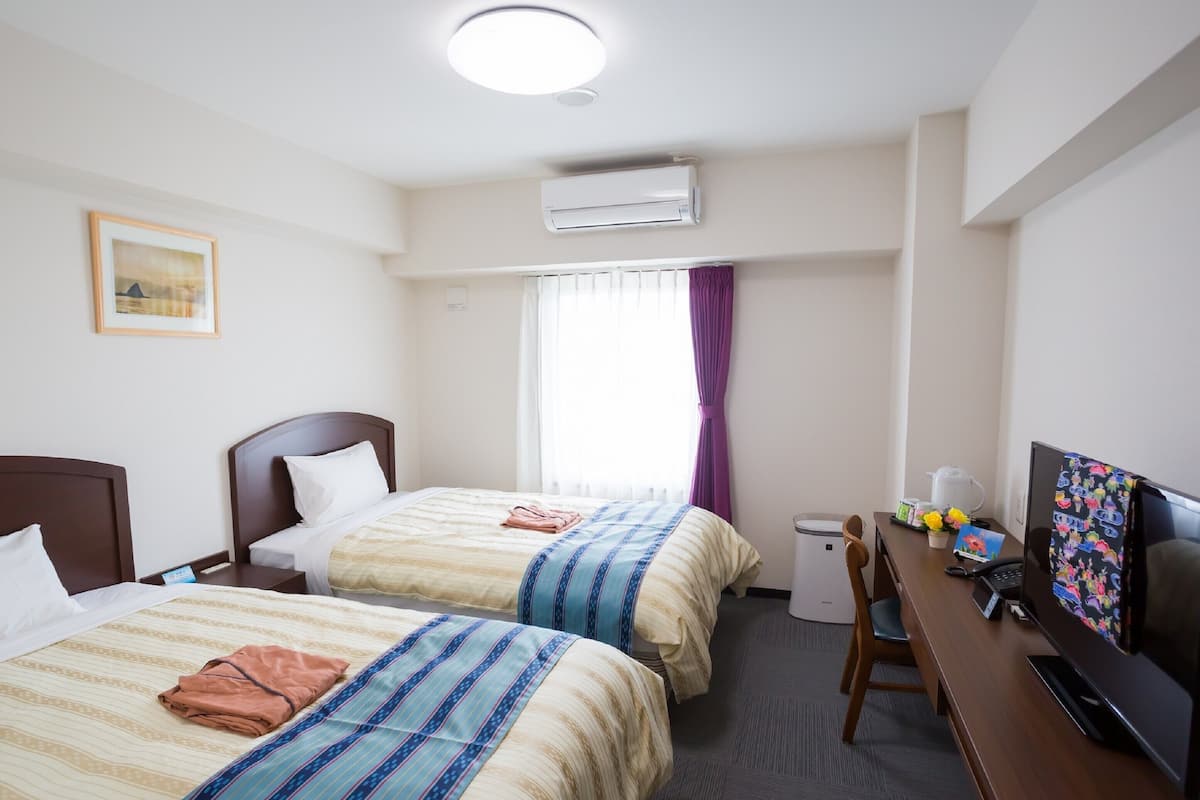 A hotel room with two beds, a desk with a computer and a TV, an air conditioning unit, and a window with curtains.