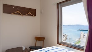 Habitación de hotel con cama, una silla, una mesita con una taza y ventana con vista al mar y un muelle.
