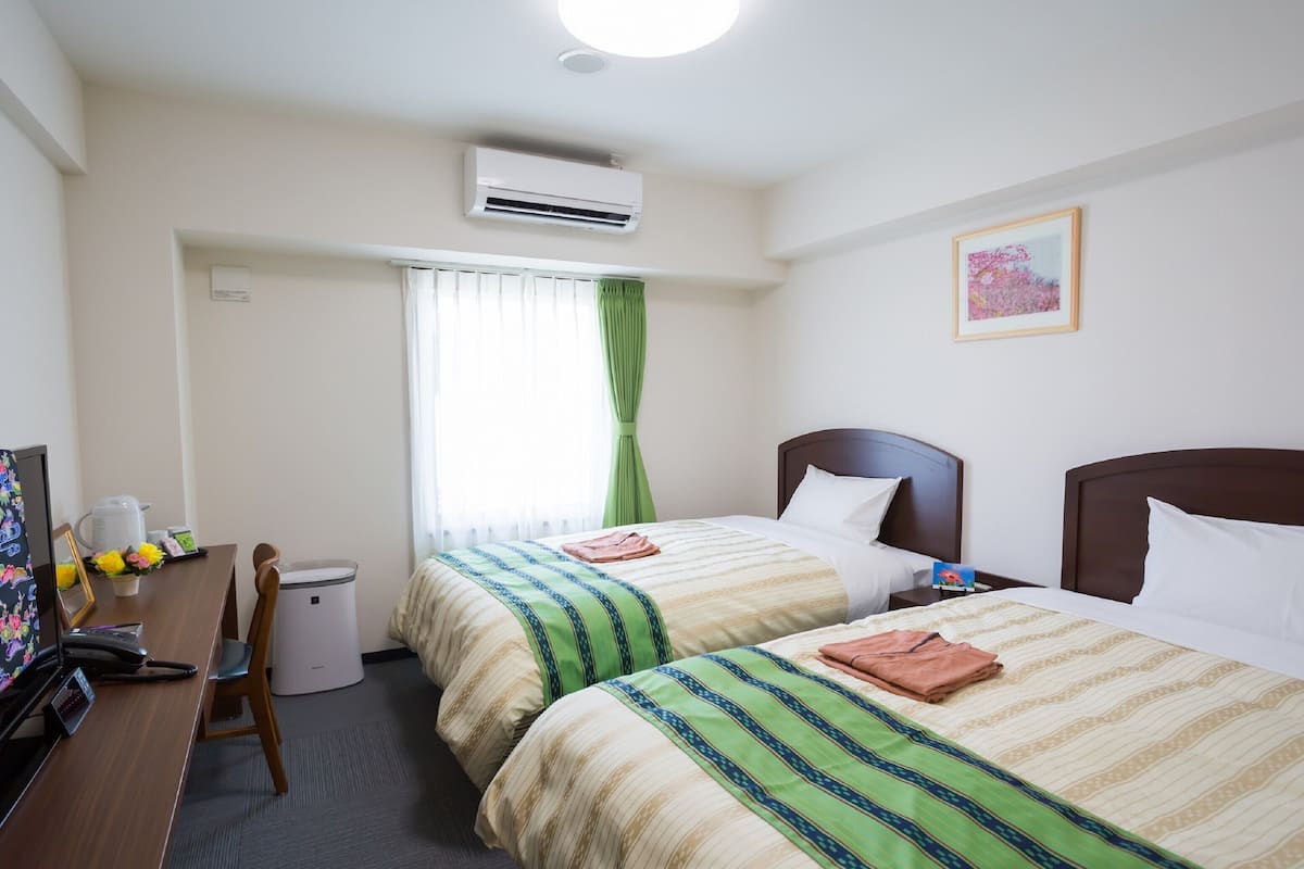 A hotel room with two beds, a desk with a telephone, a chair, a waste bin, and a window with curtains.