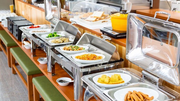 Montaje de bufé con diversos alimentos en recipientes transparentes, una barra de madera y bancos acolchados verdes.