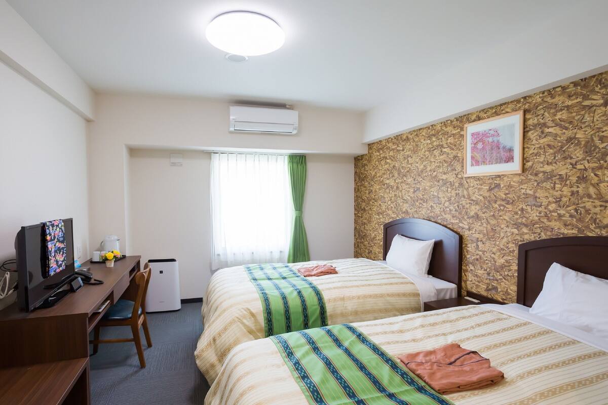 A hotel room with two beds, a desk with a computer, a small air conditioning unit, and a window with curtains.