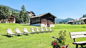 Eine grüne Rasenfläche mit Liegestühlen und einer Holzbank, ein rustikales Gebäude und ein Berg im Hintergrund.