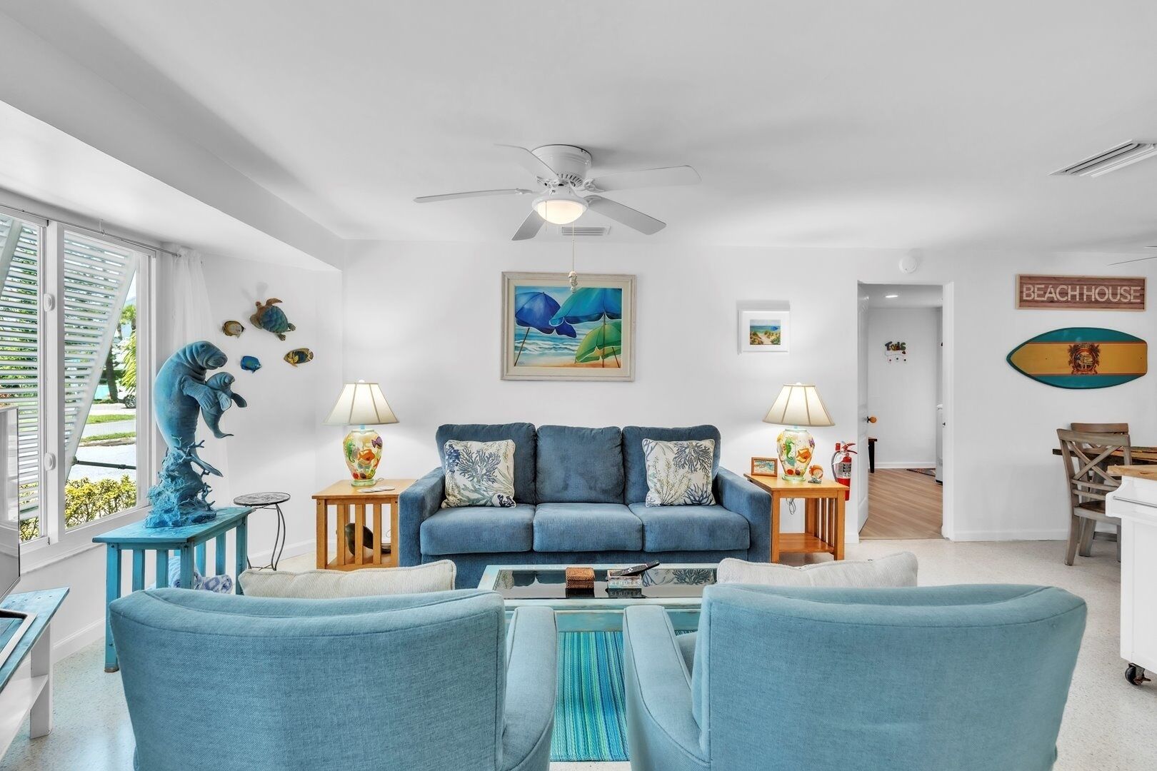 A living room with a blue sofa, two side tables with lamps, a ceiling fan, a painting of a beach scene, and a sign that says "BEACHHOUSE".