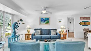 A living room with a blue sofa, two side tables with lamps, a ceiling fan, a painting of a beach scene, and a sign that says "BEACHHOUSE".