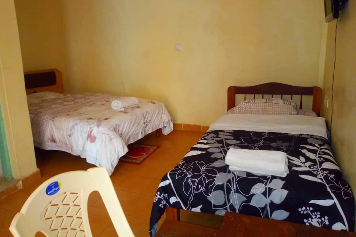 A small, simple room with two single beds, a wooden headboard, a white plastic chair, and a small table.