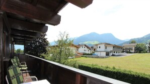 Un balcon avec une table et des chaises, donnant sur un espace verdoyant, avec des maisons et des montagnes en arrière-plan.
