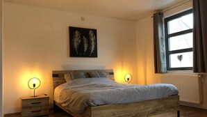 Quarto com cama de madeira, duas mesas de cabeceira com abajures, radiador e janela com cortinas.