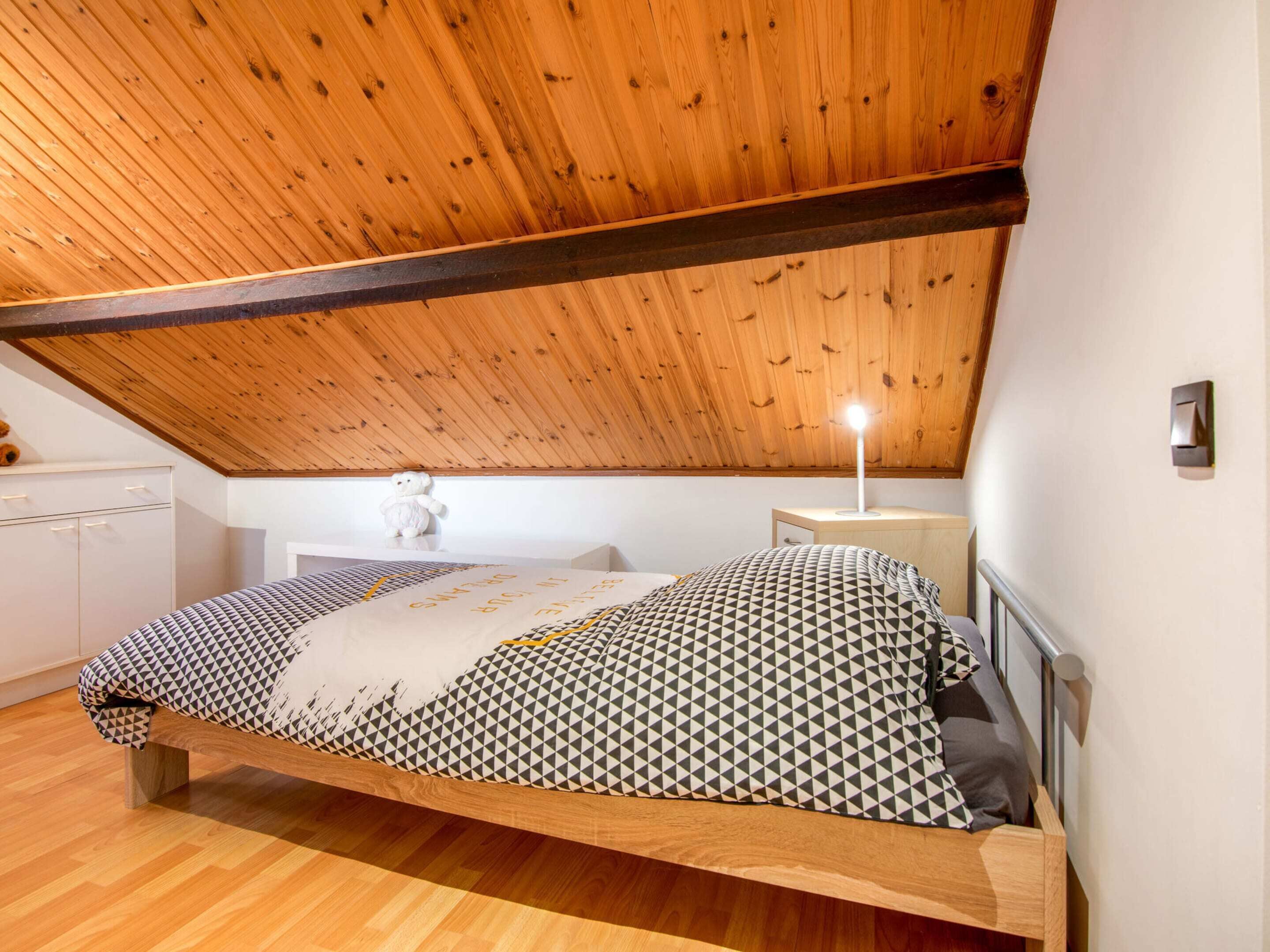 Un dormitorio con techo de madera, una cama con ropa de cama estampada, una mesita de noche y un pequeño aparador blanco.