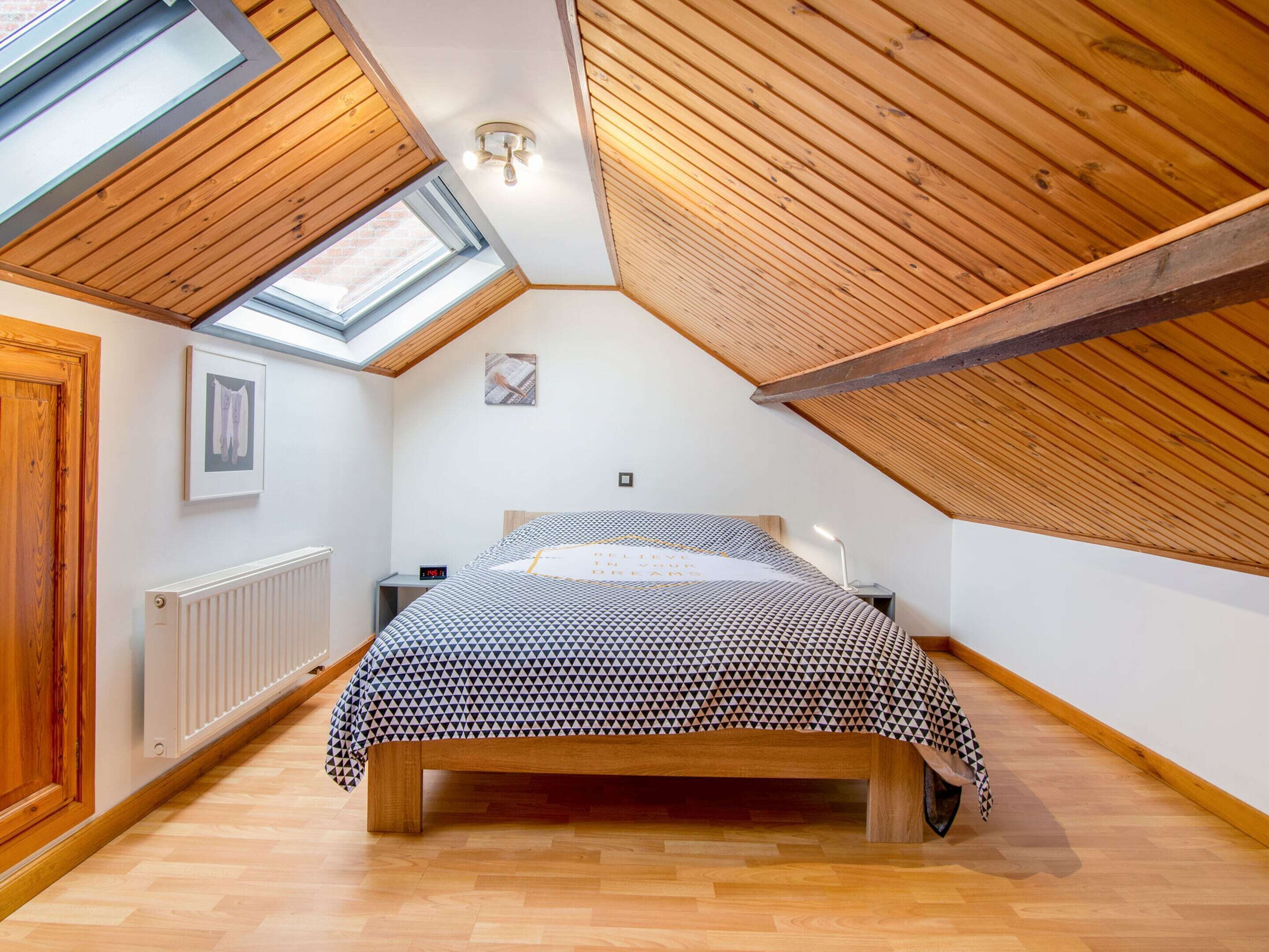 A bedroom with a sloped ceiling, wooden beams, a bed with a patterned bedspread, a bedside table with a lamp, and a window.