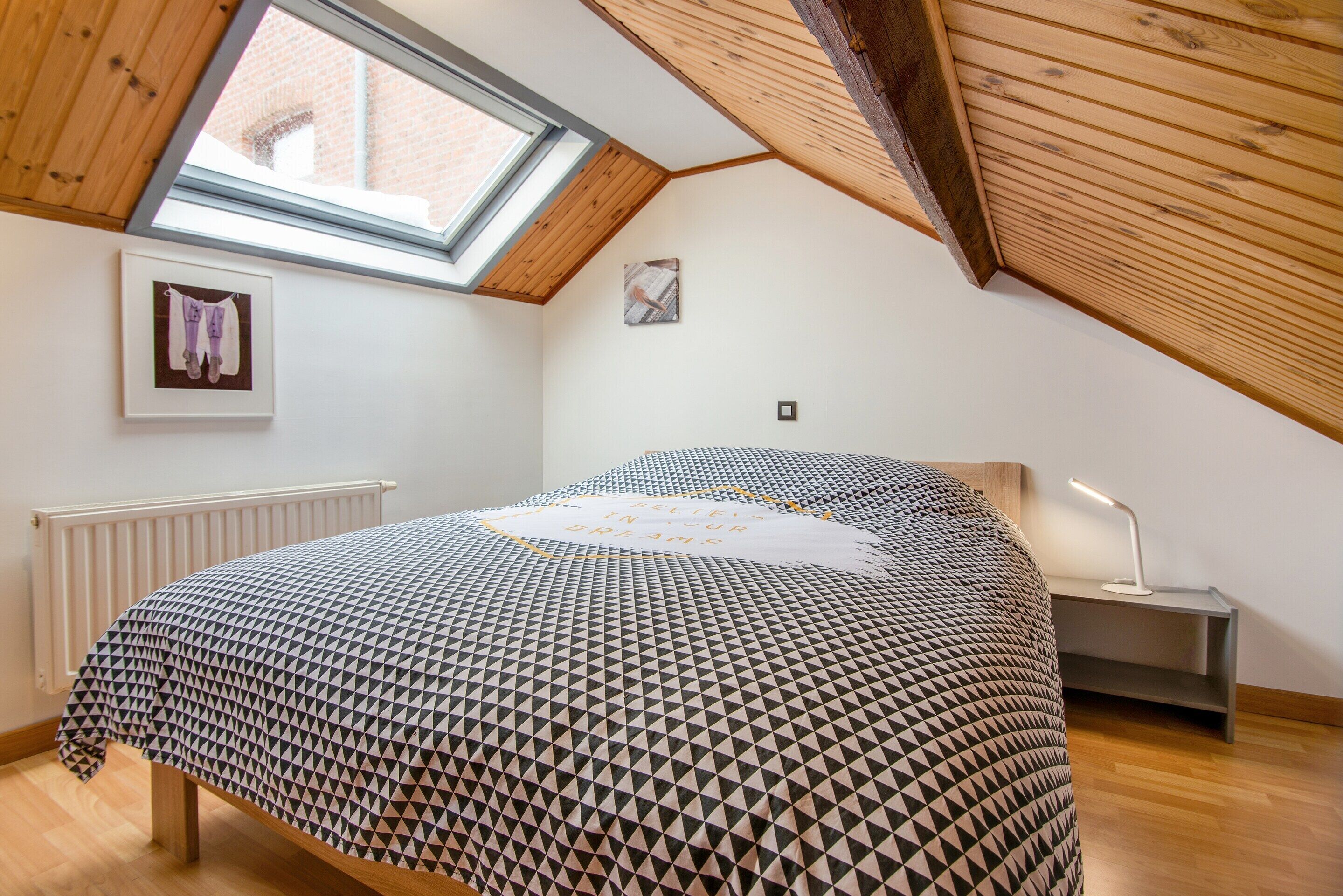 A bedroom with a sloped ceiling, a skylight, a bed with a patterned bedspread, a bedside table with a lamp, and a radiator.