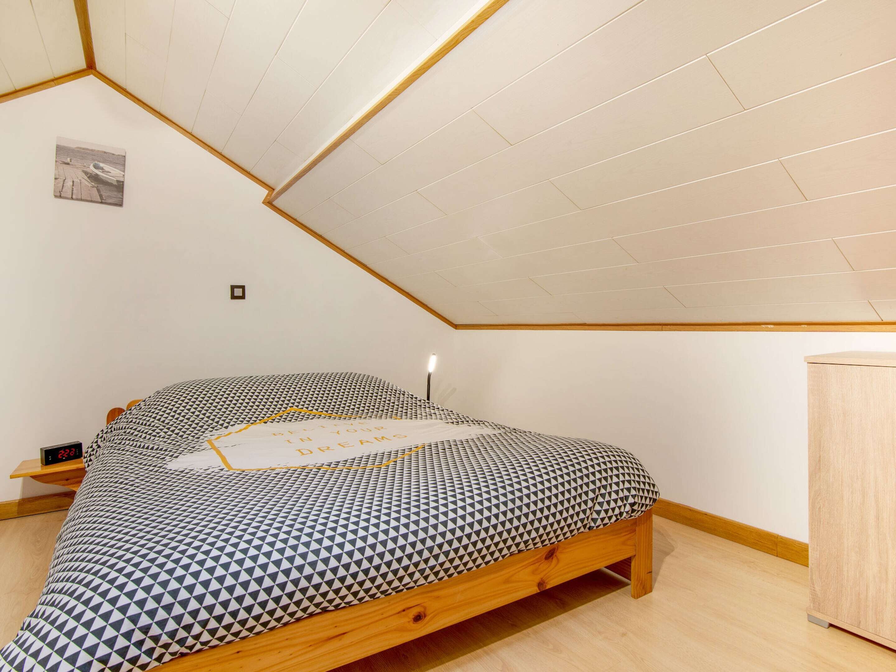 Una camera da letto con soffitto a falde, un letto con copriletto a motivi geometrici, un comodino e un tavolino notte con una sveglia.