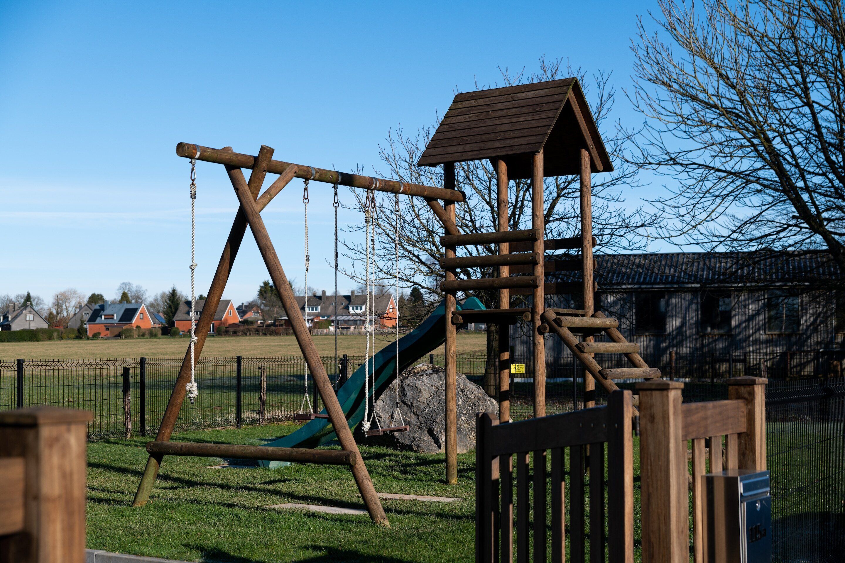 A wooden playground structure with swings and a slide, surrounded by a fenced area.